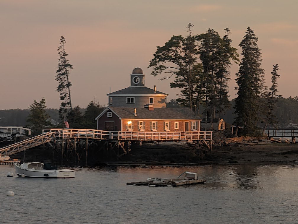 A cupola-topped boathouse glowing on a rocky point at dusk across Boothbay Harbor, a lobster boat moored in front